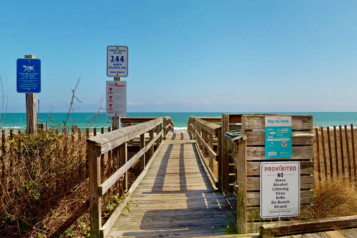 A picturesque exterior shot of a wooden boardwalk leading toward the sandy beach and ocean, with signage marking the way. The inviting pathway suggests easy access to beach activities such as walking, sunbathing, swimming, and picnicking. The ocean horizon creates a sense of adventure and possibility.