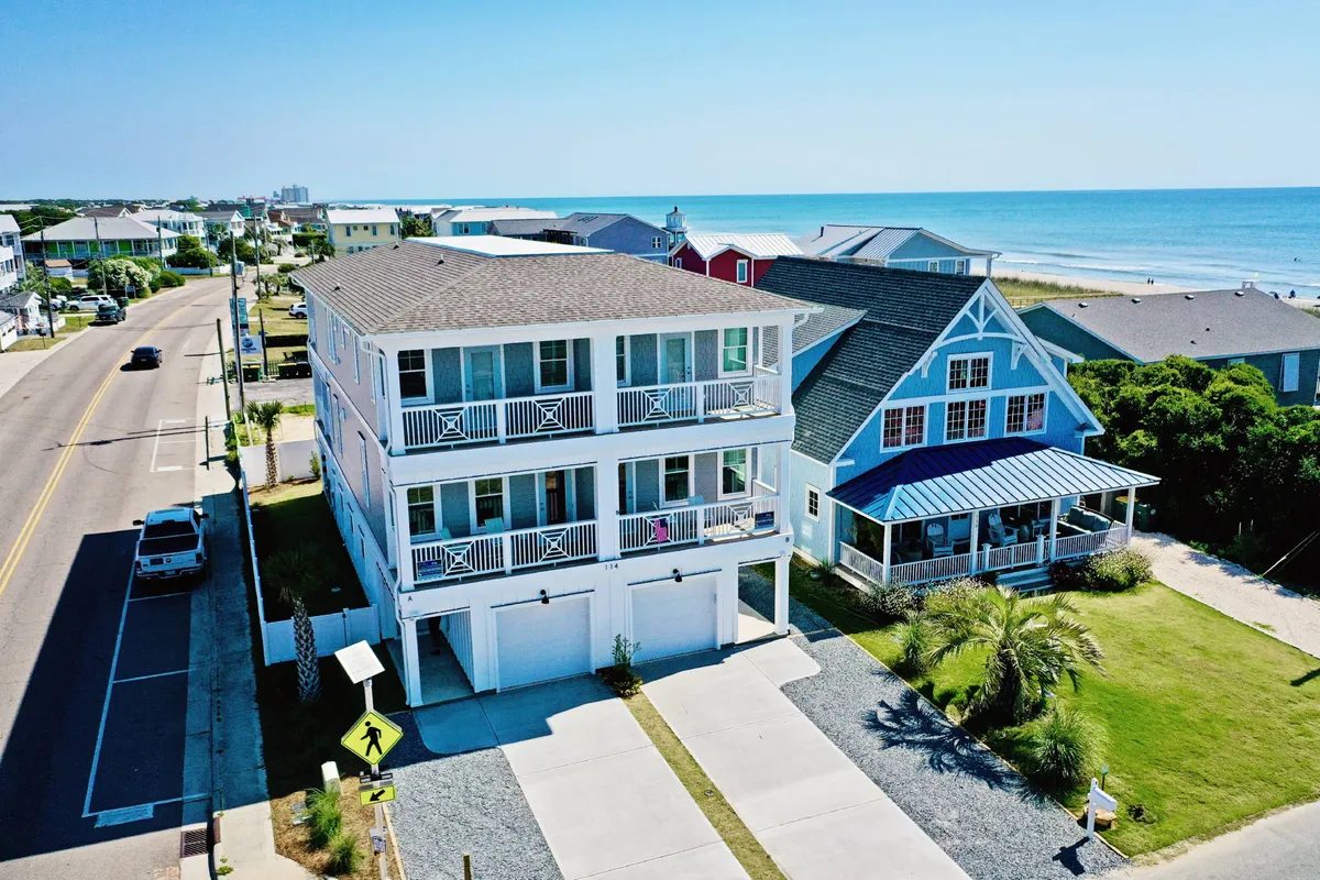 The image showcases a spacious, modern two-story beach house nestled among lush greenery, with balconies and decks offering sweeping ocean views. The house stands out in a tranquil neighborhood near Kure Beach Pier, exuding luxury and plenty of outdoor space for gatherings and relaxation.