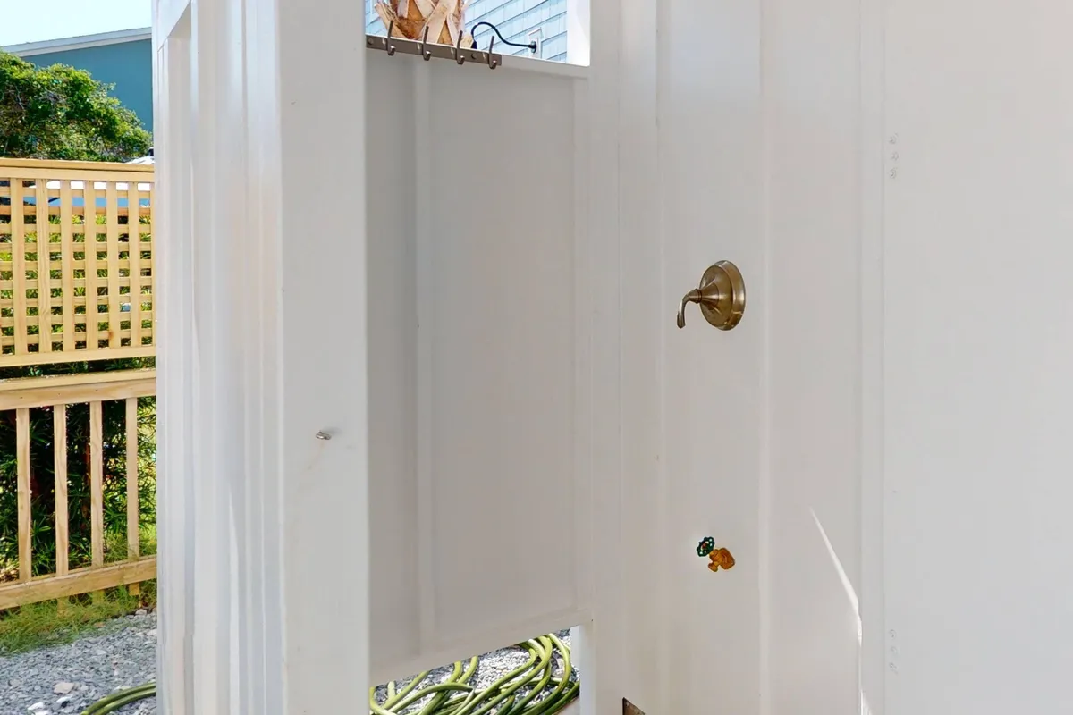 An outdoor shower space with a simple door handle, shower curtain, and hanging hooks. The design is functional and welcoming, ideal for rinsing off after a beach day before heading inside.