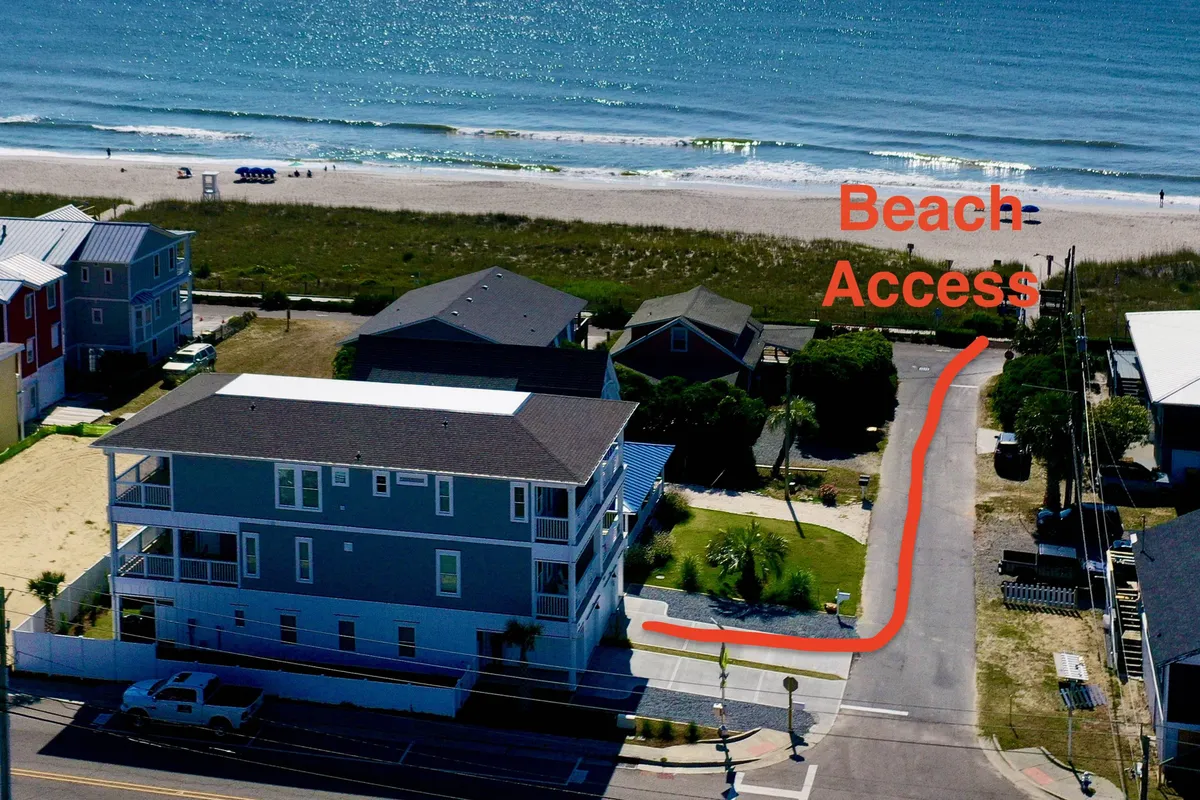 A picturesque exterior view showing a beach access pathway bordered by lush greenery, leading directly to soft sands and the expansive ocean. Framing the scene are charming coastal homes, highlighting proximity to the water and an inviting, walkable neighborhood.