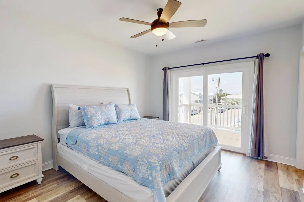 A cozy bedroom with blue patterned bedding, a ceiling fan overhead, and a sliding glass door that opens to a balcony. The room is bathed in natural light, suggesting a peaceful retreat ideal for unwinding after busy days.