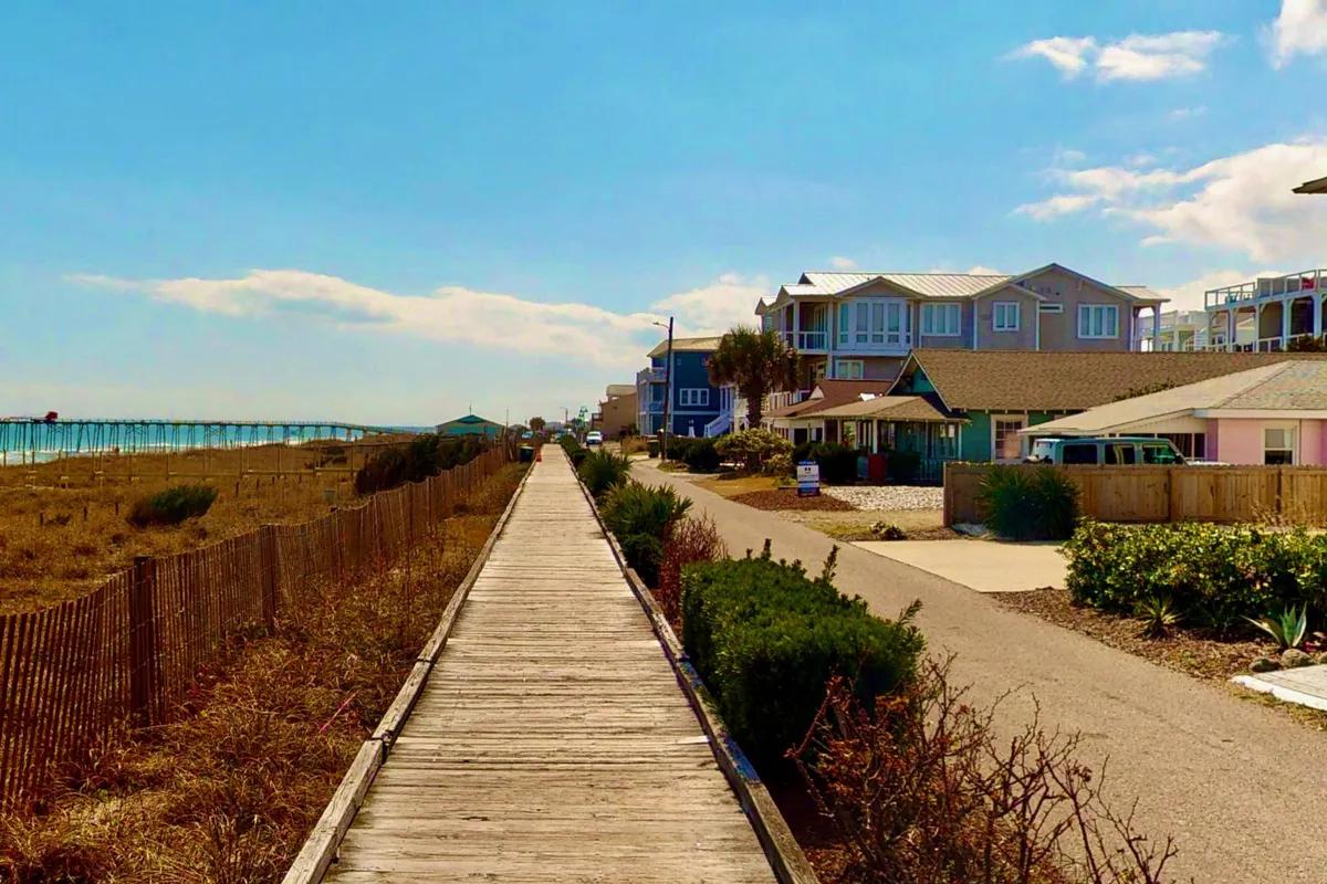 A tranquil exterior scene featuring a wooden walkway elevated above the sand, stretching toward the beach and framed by coastal homes under a bright blue sky. The view suggests a seamless transition from home to nature, ideal for leisure and relaxation.