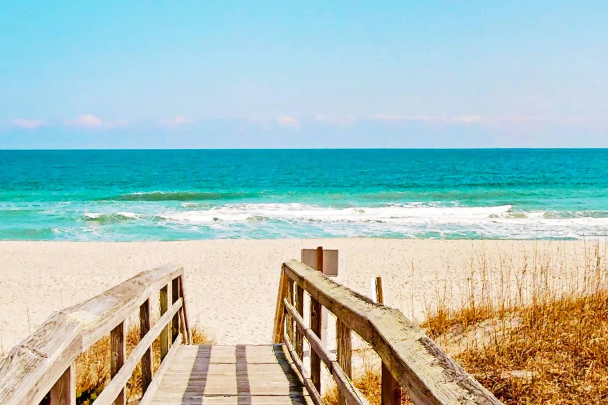 A wooden walkway leads directly over dune grass to a sandy beach with gentle ocean waves. The scene is tranquil, emphasizing direct and easy access from property to shore, with no busy road in sight. The untouched sand and natural greenery frame the walkway, inviting all ages to stroll or transport beach gear with ease.