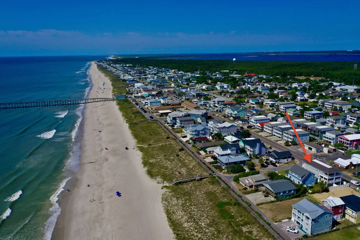 This exterior image captures the Kure Beach Pier extending out over the ocean, with nearby houses and a road visible. The beach is active but relaxed, evoking the atmosphere of morning walks, fishing, and group outings. The soft lighting suggests sunrise or sunset, emphasizing the inviting, all-day appeal of the pier and shoreline.