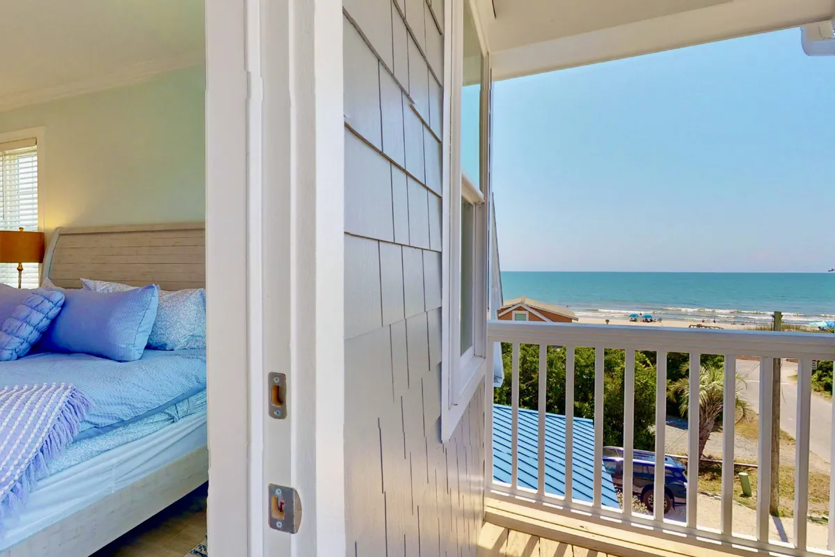 A serene bedroom featuring a comfortable bed with blue pillows, an open balcony door, and a view of the ocean just beyond. Sunlight streams through, illuminating the tranquil space and highlighting the relaxing atmosphere.