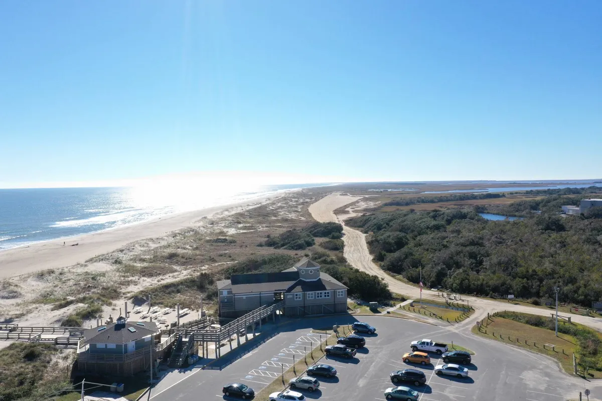 This image features a broad view of the beach and ocean, with a visible parking area and adjacent nature trails. The scene shows people engaging in beach walking and fishing, surrounded by natural coastal beauty. The tranquil atmosphere captures the essence of outdoor recreation and relaxation.