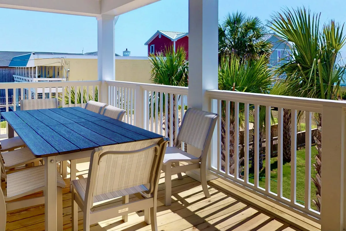 A cheerful exterior dining area with a dining table situated under palm trees, offering a comfortable space for outdoor meals or socializing. The view includes glimpses of neighboring properties, creating an inviting communal atmosphere ideal for group gatherings.
