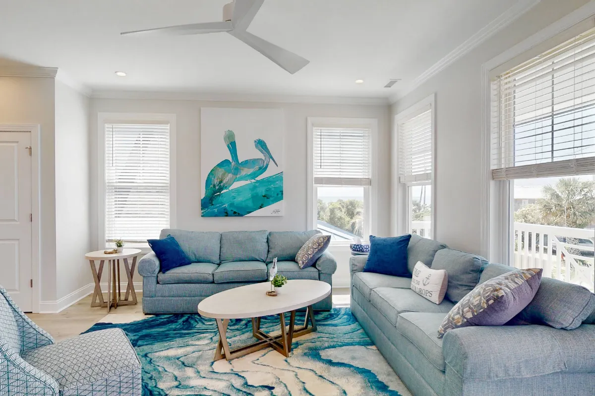 The image captures a spacious living area with light-colored sofas, a turquoise and white rug, decorative pillows, and vibrant blue artwork. The space is bathed in natural light, inviting relaxation and socialization.