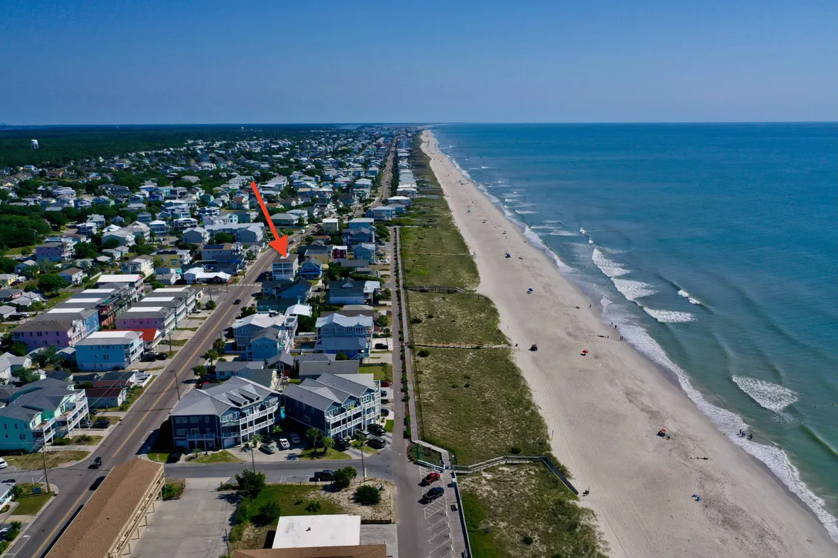 A sweeping view of the beach and ocean, with a vibrant residential area and road in the foreground, lined with houses. The image captures people enjoying the beach, walking along the shore, exploring local shops, and community events, all under a glowing sunset sky.