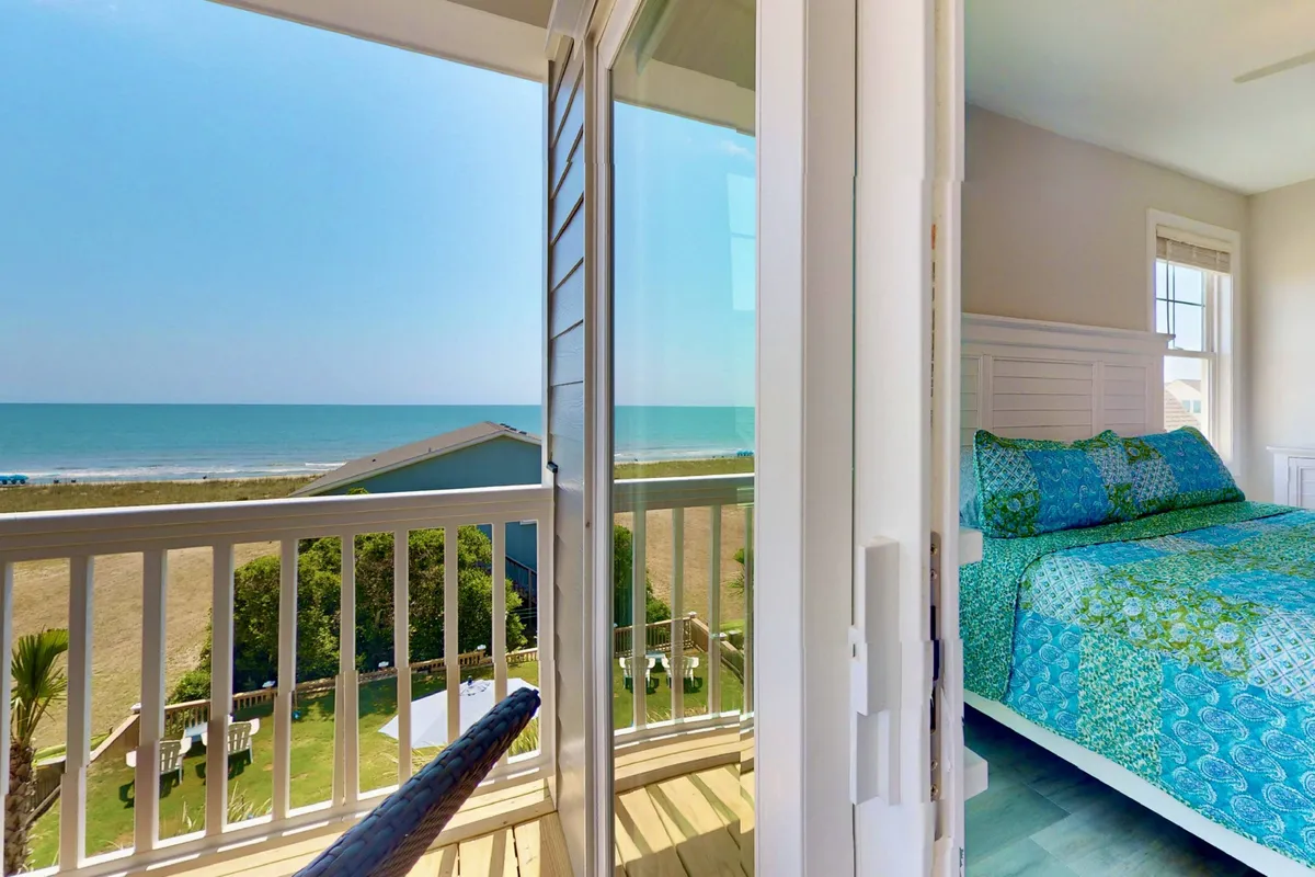 A serene view from the interior of a bedroom featuring a bed with patterned bedding, a balcony railing, and a spectacular ocean vista beyond. The scene evokes the experience of waking up to the sound and sight of the sea, blending the comfort of home with breathtaking natural beauty.